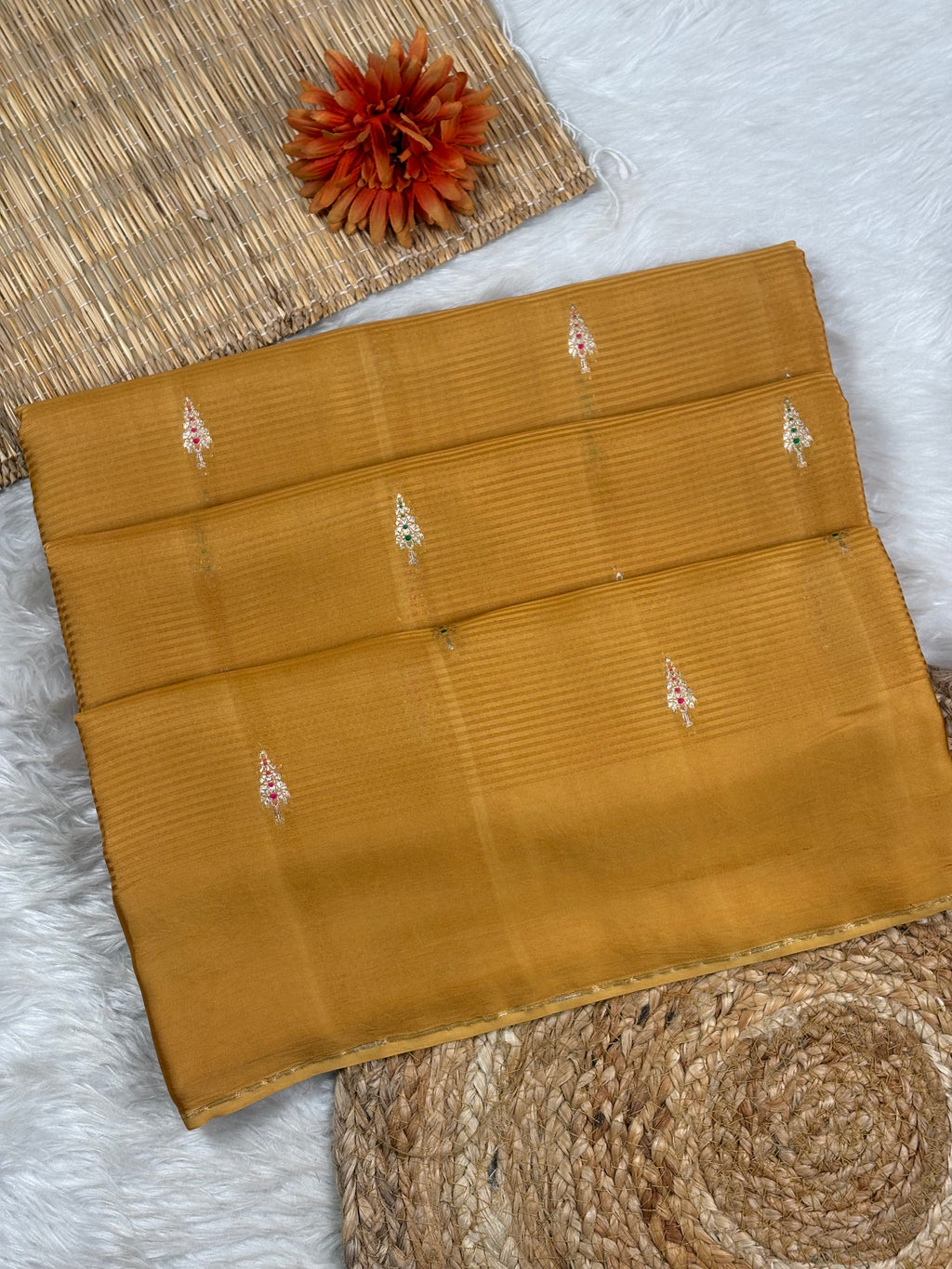 A folded mustard yellow saree with subtle horizontal pleating and small zari woven motifs, styled on a white faux-fur surface with woven mats and a dried orange flower.