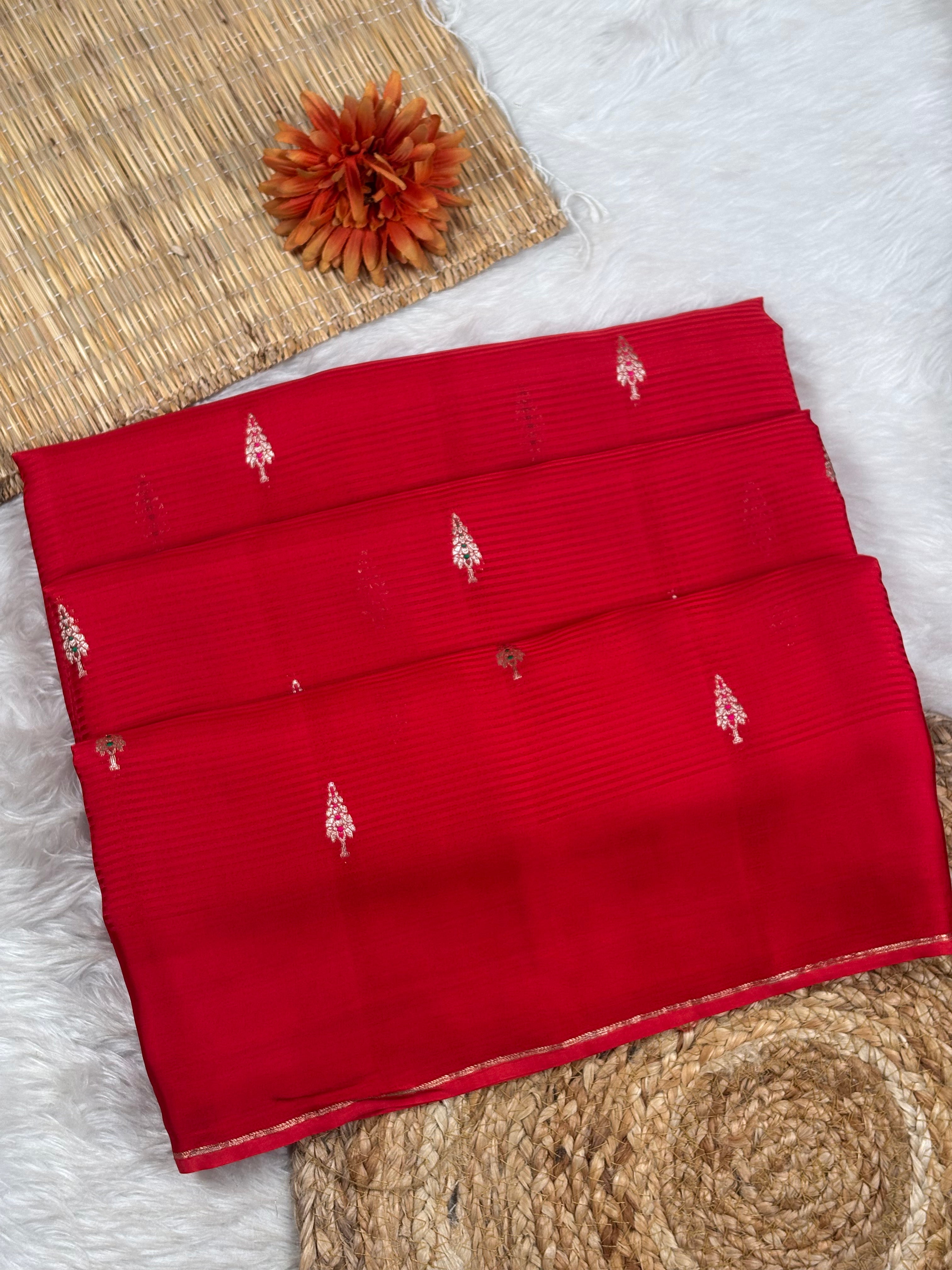 A folded red saree with small zari woven motifs, displayed on a white faux-fur surface with woven mat props.