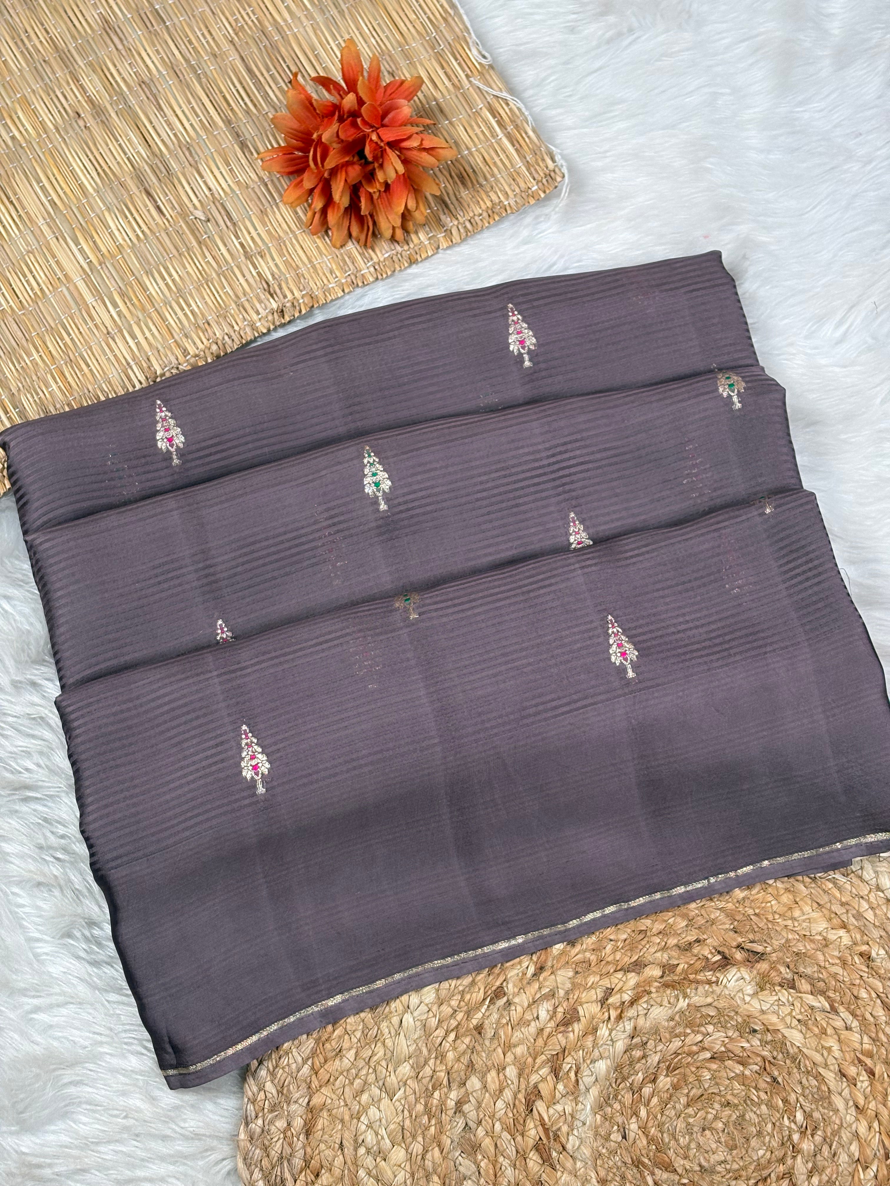 A folded dark gray-purple saree with small zari woven motifs, displayed on a white faux fur surface with a woven mat and orange flower prop.