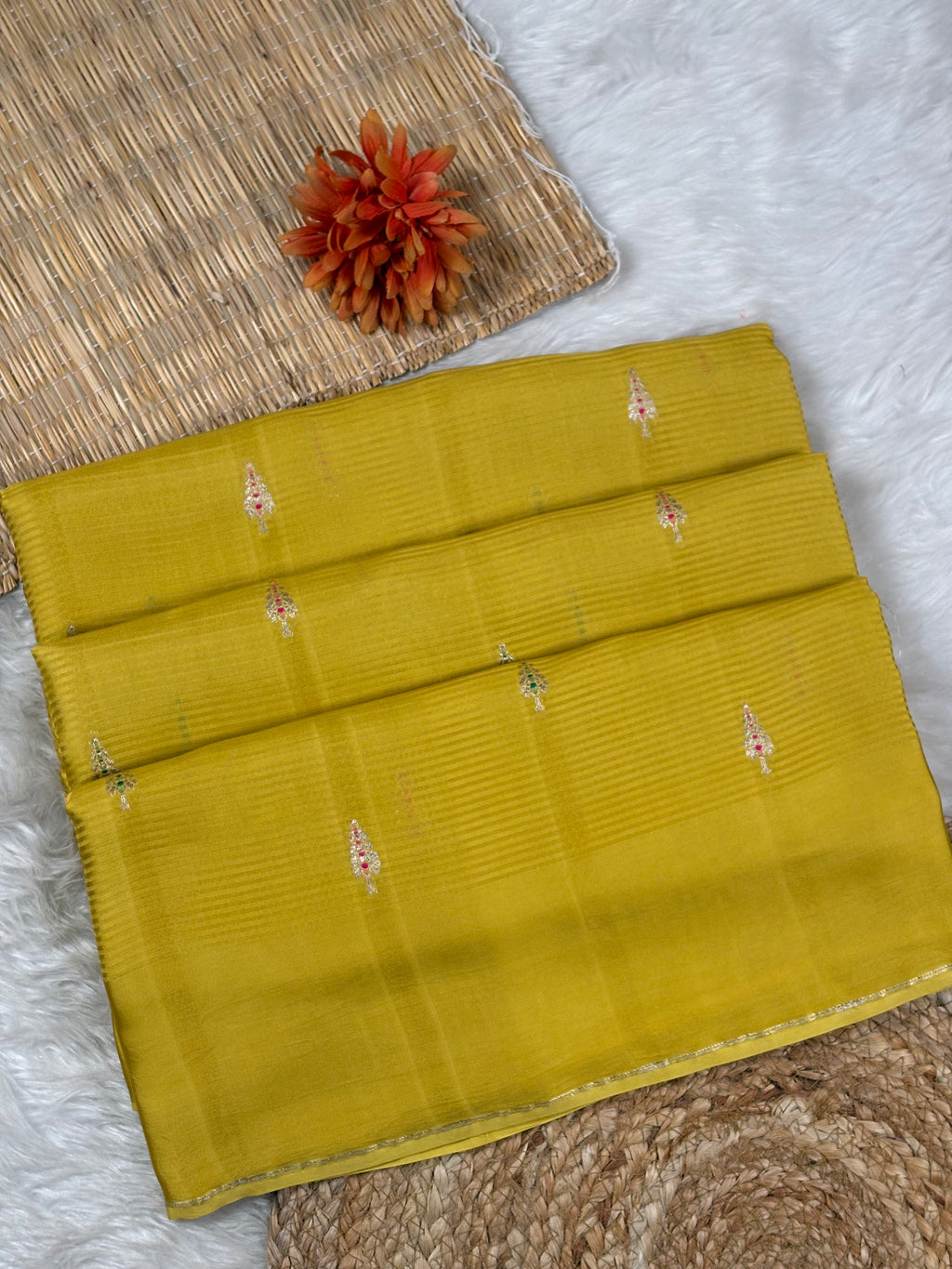 A folded yellow saree, featuring fine horizontal stripes and small zari woven motifs, displayed on a white faux fur surface with a woven mat and a decorative orange flower.