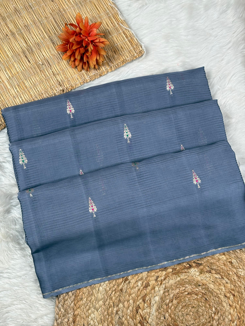 A folded slate blue saree with small zari woven motifs, displayed on a white faux fur rug with a woven mat and orange flower prop.