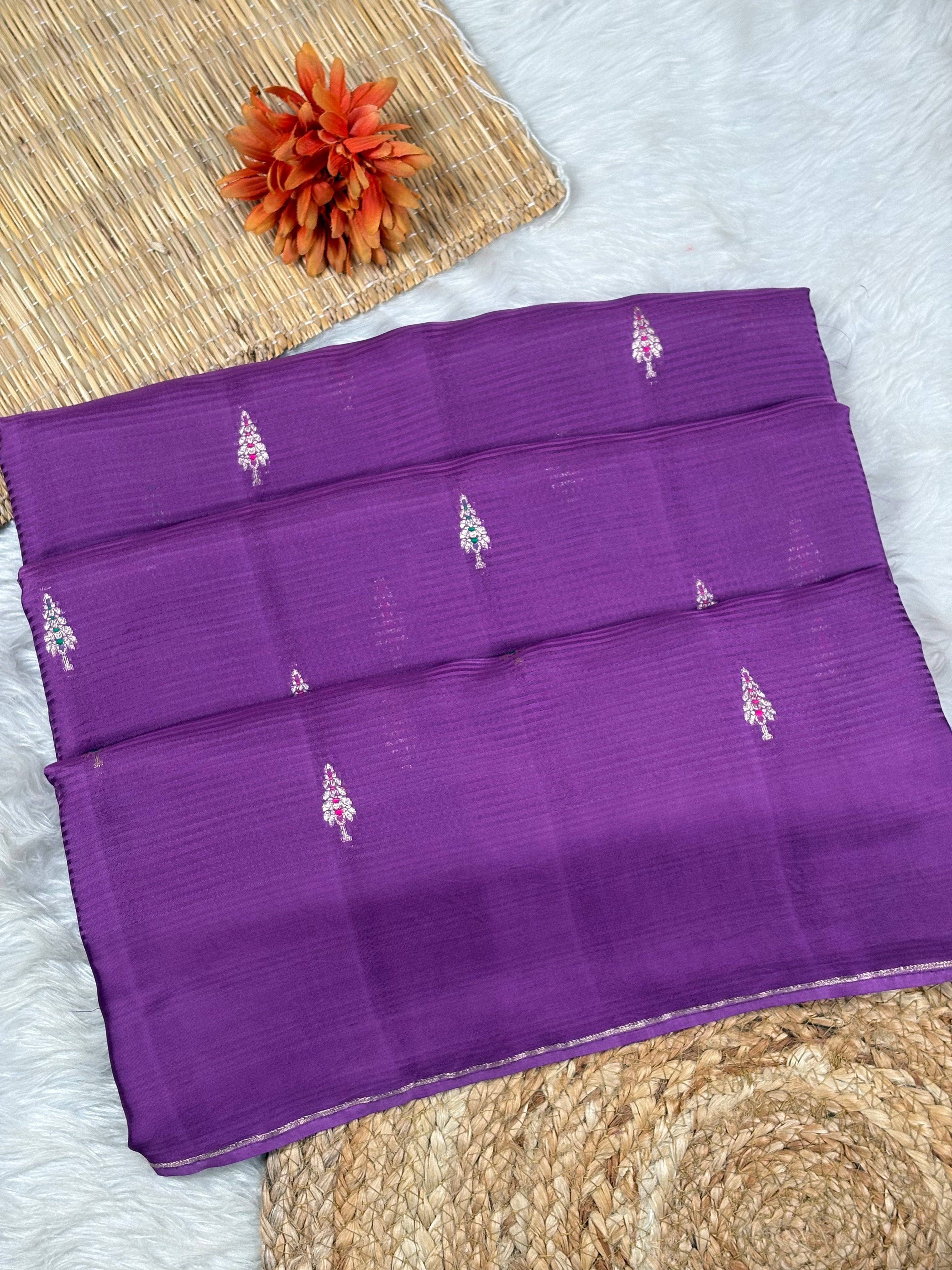 A folded purple silk saree with small zari woven motifs, displayed on a white faux fur rug with a woven mat and orange flower prop.