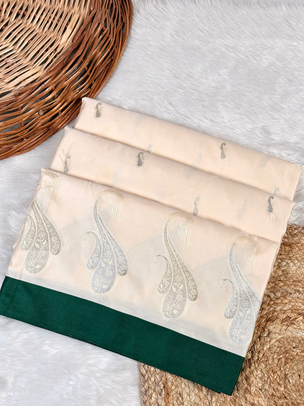 A folded cream-colored saree piece with silver paisley sona rupa weaving and a dark green border.